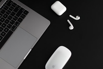 A silver laptop with a trackpad is positioned on a black surface alongside wireless earphones and their charging case. A white computer mouse is also present near the earphones.