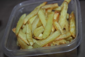 A transparent container is filled with golden-brown French fries, some of which have a crispy appearance. The container is square-shaped, and the fries are piled up in a disorganized manner.