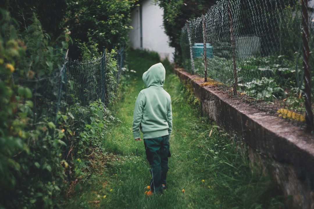 boy standing on grass and facing fence during daytime