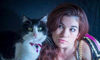 A black and white cat with striking green eyes sits beside a person with long brown hair. The cat is wearing a small red collar with a tag. The person appears contemplative, looking off to the side. The background is dark, emphasizing the subjects.