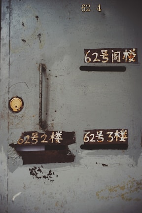 A weathered metal door with several compartments, some with Chinese characters painted above them. The paint is worn, and there are visible rust and peeling layers. A keyhole is present, and the overall condition of the door suggests age and extensive use.