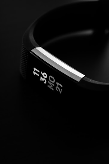 A close-up view of a digital wristwatch displaying the time and date on its screen. The watch has a sleek, modern design with a black band and metallic accents, set against a dark background.