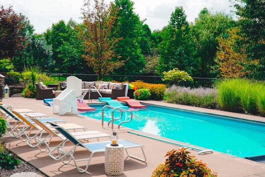 A tranquil backyard pool setting features several lounge chairs alongside a swimming pool with a slide. Cushioned patio furniture is arranged near lush greenery and colorful shrubs. The scene is accentuated with potted plants and a serene garden background.