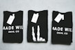 Three black T-shirts are neatly folded and laid out on a light-colored surface. The shirts have tags attached to them, and the phrase 'MADE WILD DNVR, CO' is visible on the two outer shirts, while the middle shirt features a unique white design resembling a skeleton hand.