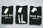 Three black T-shirts are neatly folded and laid out on a light-colored surface. The shirts have tags attached to them, and the phrase 'MADE WILD DNVR, CO' is visible on the two outer shirts, while the middle shirt features a unique white design resembling a skeleton hand.