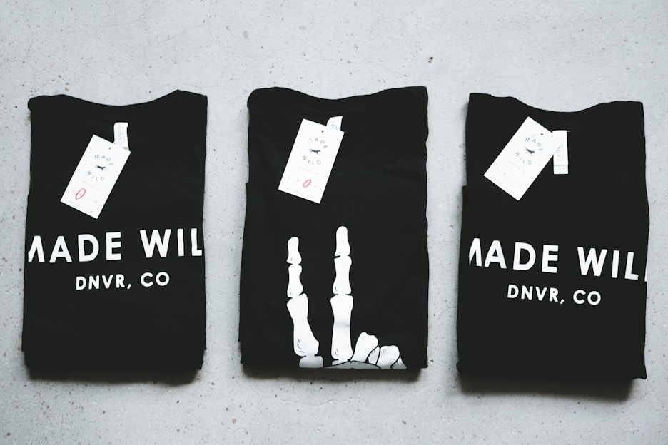 Three black T-shirts are neatly folded and laid out on a light-colored surface. The shirts have tags attached to them, and the phrase 'MADE WILD DNVR, CO' is visible on the two outer shirts, while the middle shirt features a unique white design resembling a skeleton hand.
