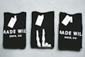 Three black T-shirts are neatly folded and laid out on a light-colored surface. The shirts have tags attached to them, and the phrase 'MADE WILD DNVR, CO' is visible on the two outer shirts, while the middle shirt features a unique white design resembling a skeleton hand.