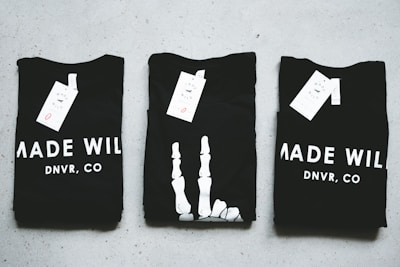 Three black T-shirts are neatly folded and laid out on a light-colored surface. The shirts have tags attached to them, and the phrase 'MADE WILD DNVR, CO' is visible on the two outer shirts, while the middle shirt features a unique white design resembling a skeleton hand.
