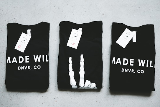 Three black T-shirts are neatly folded and laid out on a light-colored surface. The shirts have tags attached to them, and the phrase 'MADE WILD DNVR, CO' is visible on the two outer shirts, while the middle shirt features a unique white design resembling a skeleton hand.
