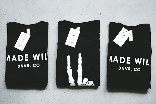 Three black T-shirts are neatly folded and laid out on a light-colored surface. The shirts have tags attached to them, and the phrase 'MADE WILD DNVR, CO' is visible on the two outer shirts, while the middle shirt features a unique white design resembling a skeleton hand.