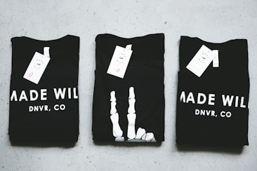 Three black T-shirts are neatly folded and laid out on a light-colored surface. The shirts have tags attached to them, and the phrase 'MADE WILD DNVR, CO' is visible on the two outer shirts, while the middle shirt features a unique white design resembling a skeleton hand.