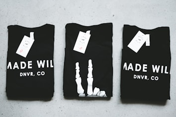 Three black T-shirts are neatly folded and laid out on a light-colored surface. The shirts have tags attached to them, and the phrase 'MADE WILD DNVR, CO' is visible on the two outer shirts, while the middle shirt features a unique white design resembling a skeleton hand.