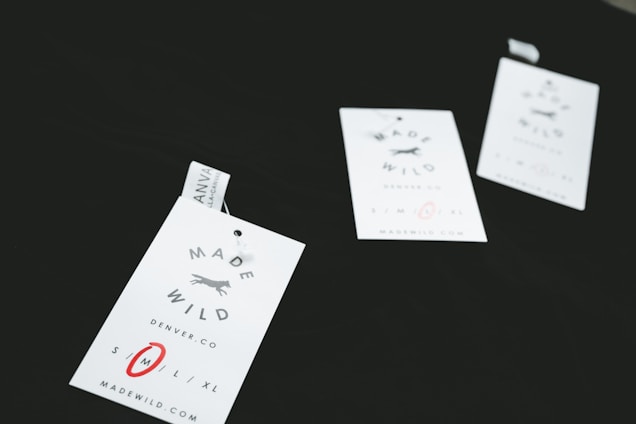Three clothing tags with the words 'Made Wild' and 'Denver, CO' are arranged on a dark surface. The tags include size options with 'M' circled in red. Each tag has a small illustration of a running fox.