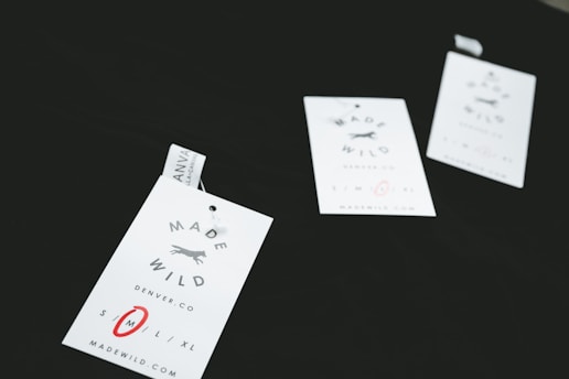 Three clothing tags with the words 'Made Wild' and 'Denver, CO' are arranged on a dark surface. The tags include size options with 'M' circled in red. Each tag has a small illustration of a running fox.