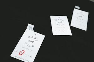 Three clothing tags with the words 'Made Wild' and 'Denver, CO' are arranged on a dark surface. The tags include size options with 'M' circled in red. Each tag has a small illustration of a running fox.