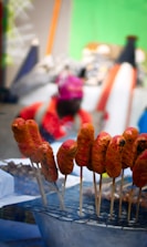 Skewered sausages coated in a reddish-orange marinade are displayed on a grill. The background shows an unfocused person wearing a bright pink headscarf and red clothing, suggesting a vibrant street market or outdoor setting.