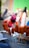 Skewered sausages coated in a reddish-orange marinade are displayed on a grill. The background shows an unfocused person wearing a bright pink headscarf and red clothing, suggesting a vibrant street market or outdoor setting.