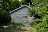 A weathered garage with a gabled roof, partially obscured by dense green foliage. The garage door has a 'No Parking' sign and appears aged, with patches of peeling paint.