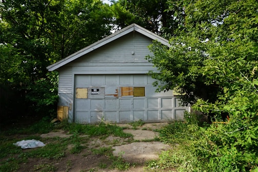 A weathered garage with a gabled roof, partially obscured by dense green foliage. The garage door has a 'No Parking' sign and appears aged, with patches of peeling paint.