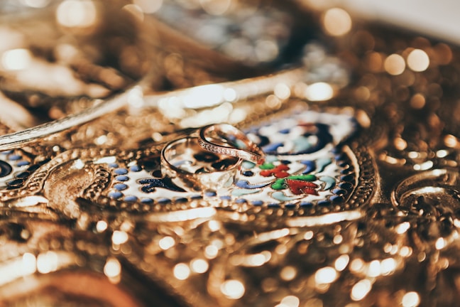 Intricate gold jewelry serves as a detailed background with ornate engravings, featuring two elegant gold rings placed prominently at the center.