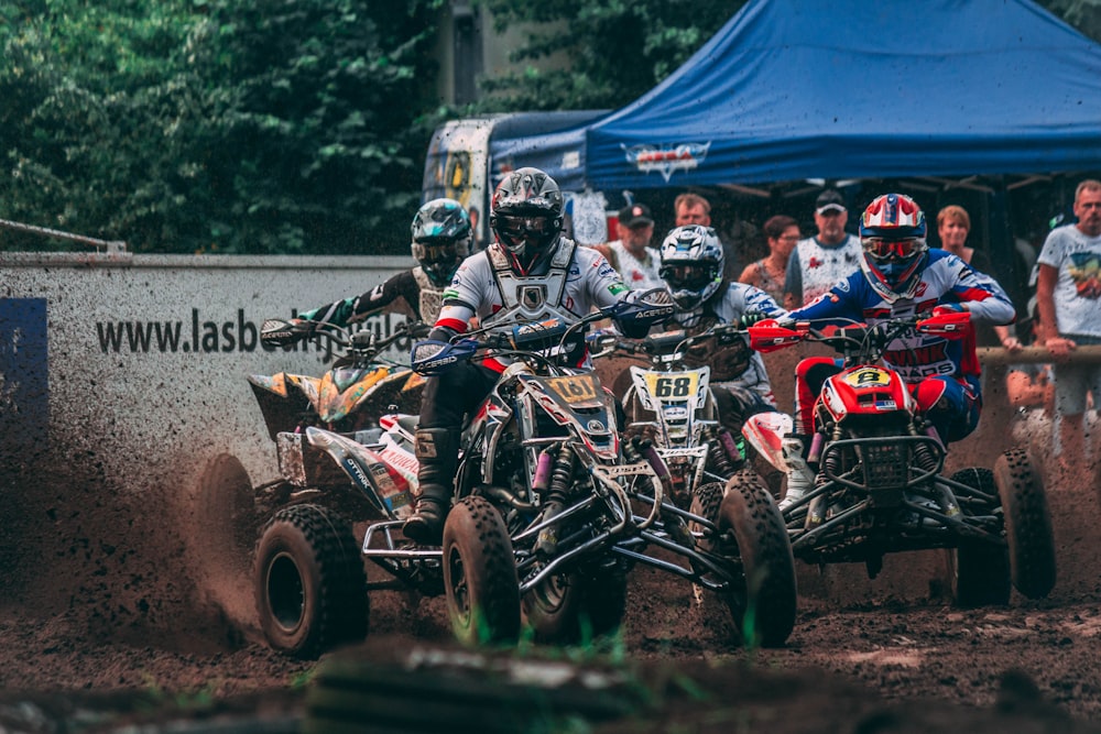 four men riding ATVs racing on mud with people watching during daytime