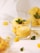 A creamy yellow dessert served in a small glass, garnished with fresh mint leaves and surrounded by chunks of mango. The setting includes a white background with additional glasses of dessert in the background, creating a fresh and inviting presentation.