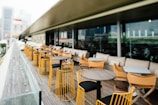 A modern outdoor patio with rows of tables and chairs made of wood and metal, featuring yellow and black accents. The area is designed for dining, with beige cushions lining the seats. Glass panels run along the edge, offering a view of the cityscape beyond.