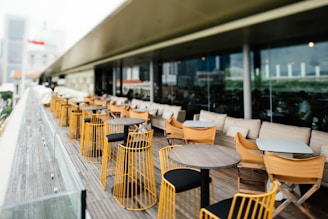 A modern outdoor patio with rows of tables and chairs made of wood and metal, featuring yellow and black accents. The area is designed for dining, with beige cushions lining the seats. Glass panels run along the edge, offering a view of the cityscape beyond.