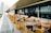 A modern outdoor patio with rows of tables and chairs made of wood and metal, featuring yellow and black accents. The area is designed for dining, with beige cushions lining the seats. Glass panels run along the edge, offering a view of the cityscape beyond.