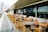 A modern outdoor patio with rows of tables and chairs made of wood and metal, featuring yellow and black accents. The area is designed for dining, with beige cushions lining the seats. Glass panels run along the edge, offering a view of the cityscape beyond.