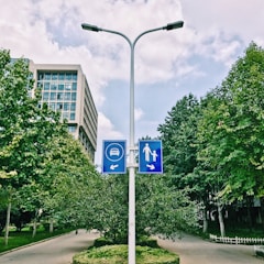 A street lamp post with two blue traffic signs indicating directions for vehicles to the left and pedestrian path to the right. The background consists of a modern office building, lush green trees, and clear skies with scattered clouds.