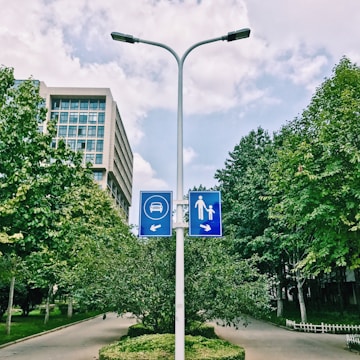 A street lamp post with two blue traffic signs indicating directions for vehicles to the left and pedestrian path to the right. The background consists of a modern office building, lush green trees, and clear skies with scattered clouds.