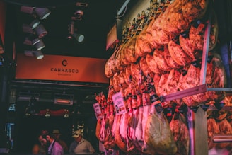 An array of cured meat legs hanging in a market stall, with warm lighting casting a reddish hue. The signage displays the brand name Carrasco, suggesting a specialty in Iberico ham. Several people in hats and casual wear are observing the display.