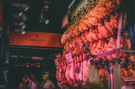 An array of cured meat legs hanging in a market stall, with warm lighting casting a reddish hue. The signage displays the brand name Carrasco, suggesting a specialty in Iberico ham. Several people in hats and casual wear are observing the display.