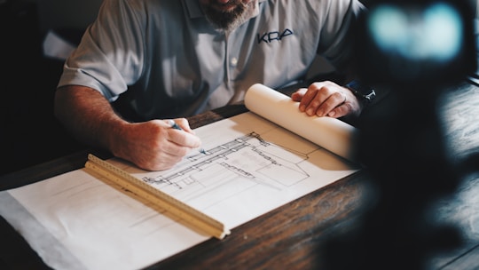 A person is seated at a desk, working on architectural plans. They are using a pencil to make adjustments on a large sheet of paper with detailed drawings. A ruler is placed parallel to the edge of the paper, suggesting precision work. The person's focus is evident, and they are dressed in a casual shirt.