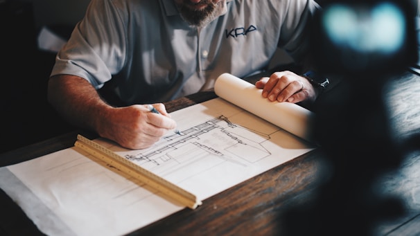 A person is seated at a desk, working on architectural plans. They are using a pencil to make adjustments on a large sheet of paper with detailed drawings. A ruler is placed parallel to the edge of the paper, suggesting precision work. The person's focus is evident, and they are dressed in a casual shirt.