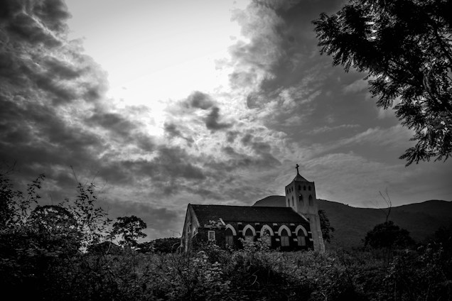 A black-and-white image of a small church with a cross on its steeple, set against a dramatic sky filled with thick clouds. The church is surrounded by dense vegetation, and a hill can be seen in the background, creating a serene yet mysterious atmosphere.