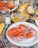 A cooked lobster is placed on a white plate surrounded by a variety of foods, including corn on the cob and glasses of drinks. The tablecloth features an intricate pattern, adding a vibrant backdrop to the meal. Condiments like salt and pepper shakers are also visible, suggesting a casual dining setting.
