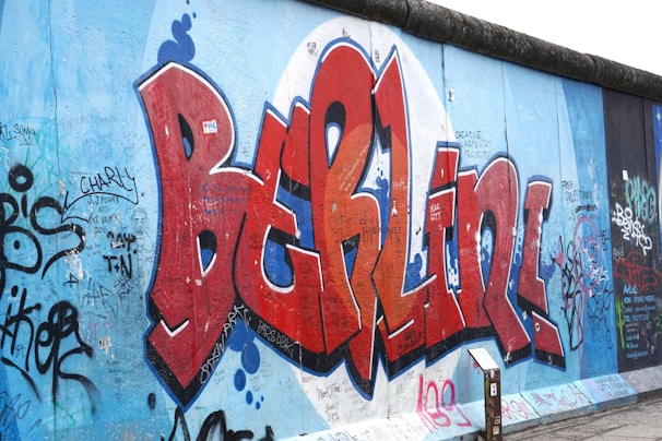 A section of a graffiti-covered wall featuring the word 'Berlin' in large, bold red letters with a blue and white background. The wall is filled with various tags, artwork, and scribbles in different colors, adding to the urban and artistic atmosphere.