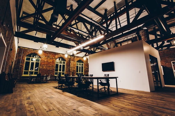 An industrial-style meeting room with exposed wooden beams and brick walls. The space is furnished with modern office chairs and tables. Large windows with arched frames line the walls, and pendant lights hang from the ceiling. The wooden floor adds warmth to the ambiance.