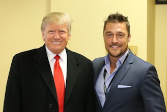 Two men are standing side by side, both smiling at the camera. One is wearing a black coat with a red tie, while the other is dressed in a blue suit with an open-collared shirt. The background is a neutral beige color, creating a professional atmosphere.
