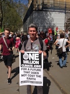 A person stands at a demonstration holding a large sign that advocates for climate jobs. The sign says 'One Million Climate Jobs for a Future that Does Not Cost the Earth' with a link to an environmental campaign website. People in the background suggest a protest or rally environment.