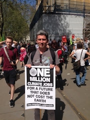A person stands at a demonstration holding a large sign that advocates for climate jobs. The sign says 'One Million Climate Jobs for a Future that Does Not Cost the Earth' with a link to an environmental campaign website. People in the background suggest a protest or rally environment.