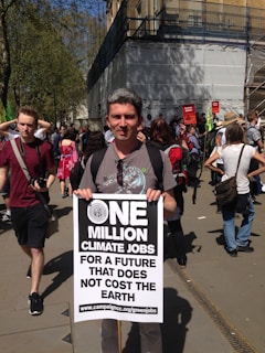 A person stands at a demonstration holding a large sign that advocates for climate jobs. The sign says 'One Million Climate Jobs for a Future that Does Not Cost the Earth' with a link to an environmental campaign website. People in the background suggest a protest or rally environment.