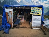 A small shop situated in a mountainous area, covered with a blue tarp. Several shelves inside the shop display various goods such as snacks and drinks. A poster with pictures is hanging near the entrance. The background features majestic mountains and a cloudy sky, creating a scenic view.