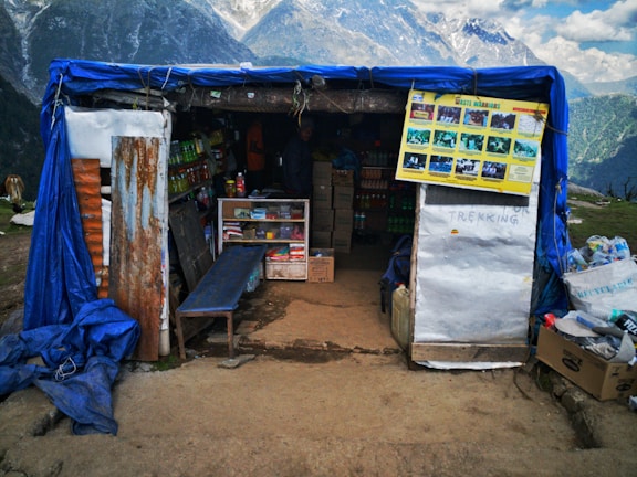 A small shop situated in a mountainous area, covered with a blue tarp. Several shelves inside the shop display various goods such as snacks and drinks. A poster with pictures is hanging near the entrance. The background features majestic mountains and a cloudy sky, creating a scenic view.