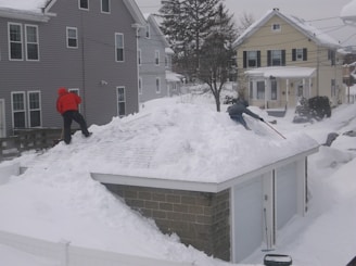 Two individuals are shoveling snow off the roof of a small garage. One person is wearing a red jacket and black pants, while the other is in a green jacket. The surrounding area is covered in snow, including nearby houses with light-colored siding. A tree without leaves stands in the background, indicating a winter setting.