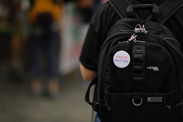 A close-up view of a black backpack with multiple compartments, featuring a button attached that reads 'FACTS MATTER' in red and blue letters.