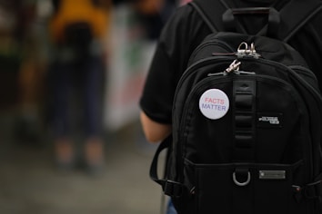 A close-up view of a black backpack with multiple compartments, featuring a button attached that reads 'FACTS MATTER' in red and blue letters.