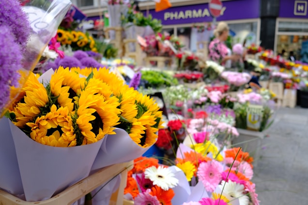 Bright and colorful sunflowers are prominently displayed alongside an assortment of other flowers like gerberas and chrysanthemums. The background reveals a bustling street flower market with a variety of floral arrangements, set against the facade of a pharmacy.